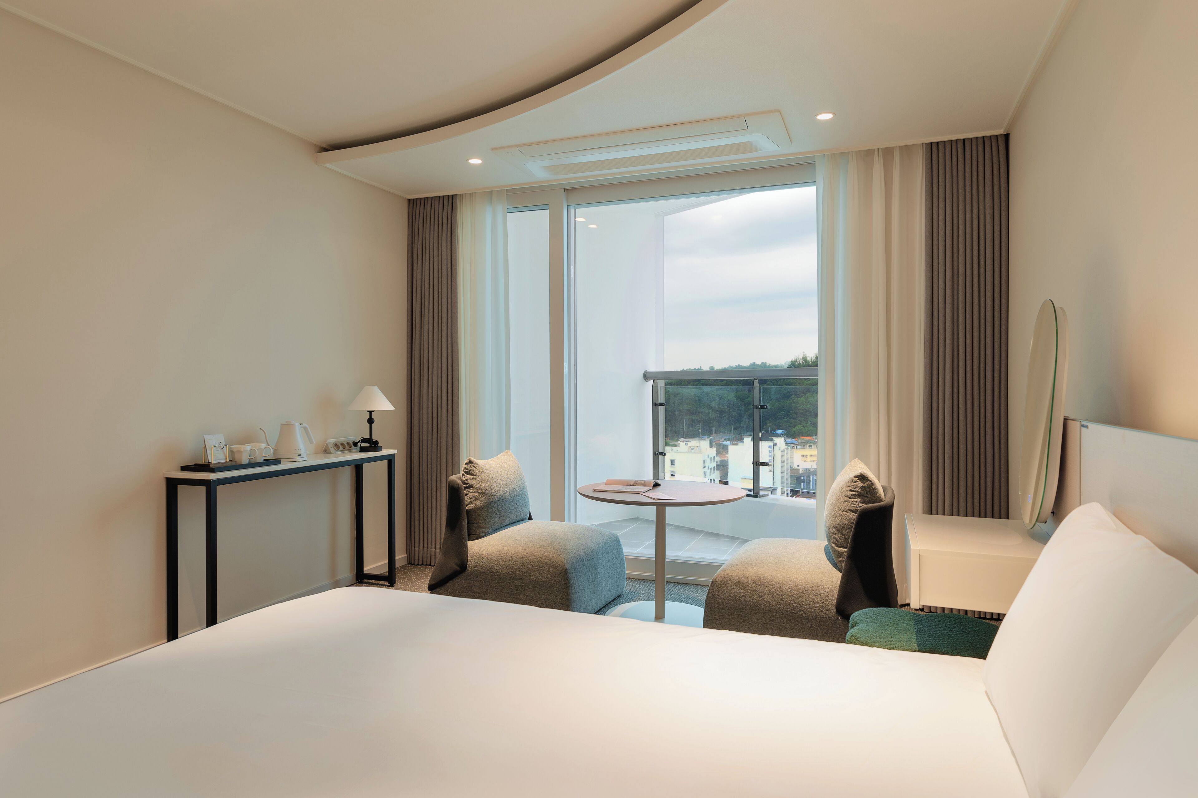 A hotel room with a large bed, a small round table, two armchairs, a desk with a lamp, and a large window offering a view of the city.