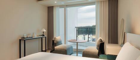 A hotel room with a large bed, a small round table, two armchairs, a desk with a lamp, and a large window offering a view of the city.