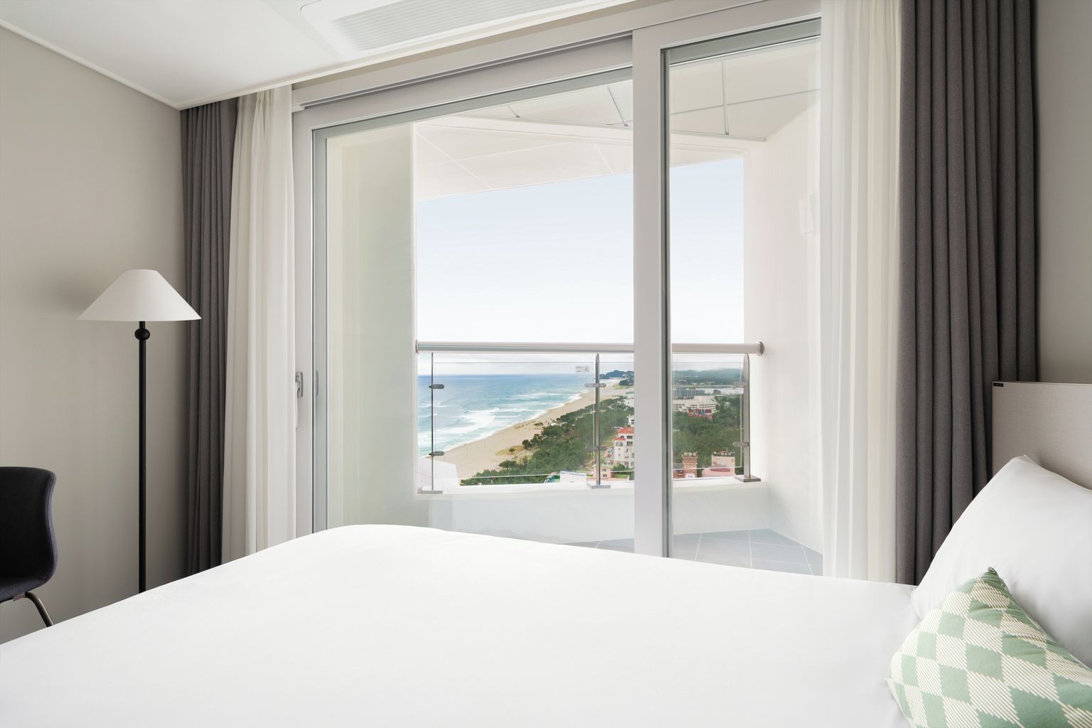 A hotel room with a bed, a chair, a lamp, and a large sliding glass door leading to a balcony with a view of the beach.