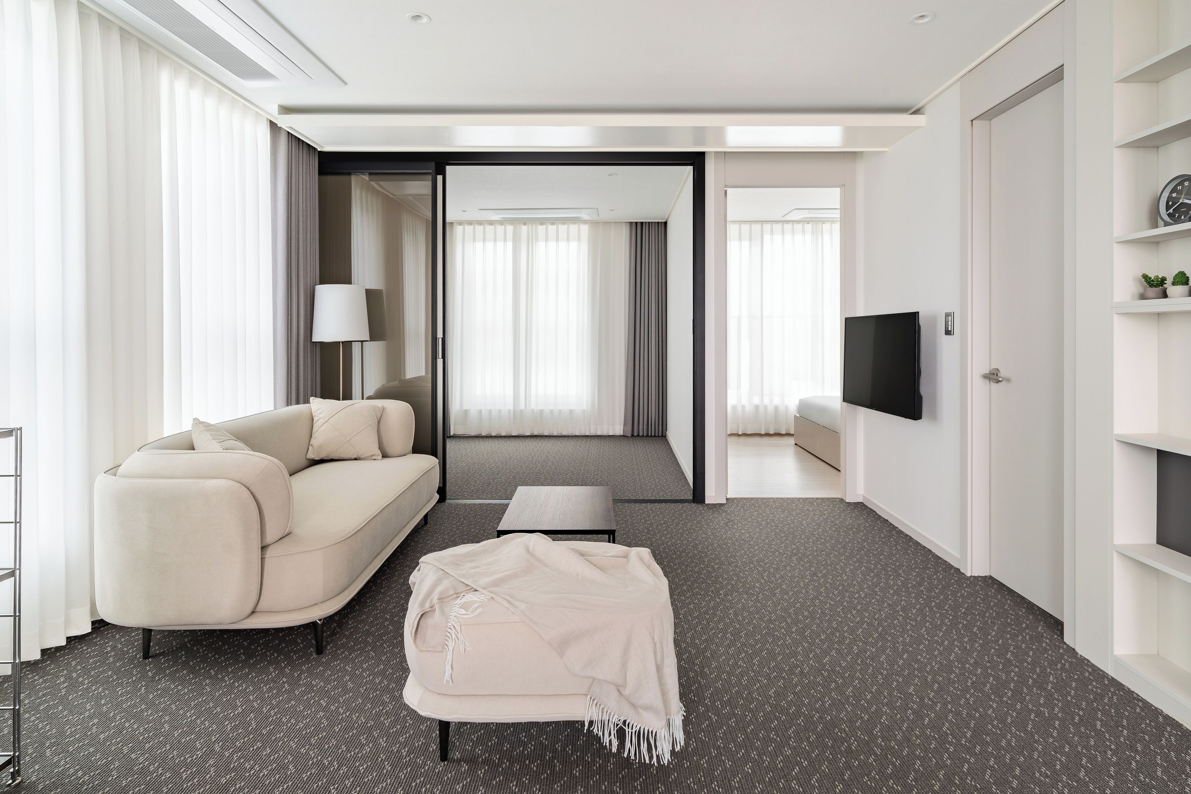 A modern hotel room with a sofa, a small table, a TV, and a large window with curtains.
