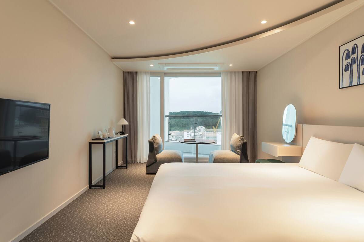 A hotel room with a large bed, a TV, a desk with a chair, a small table, and a view of the city through large windows.