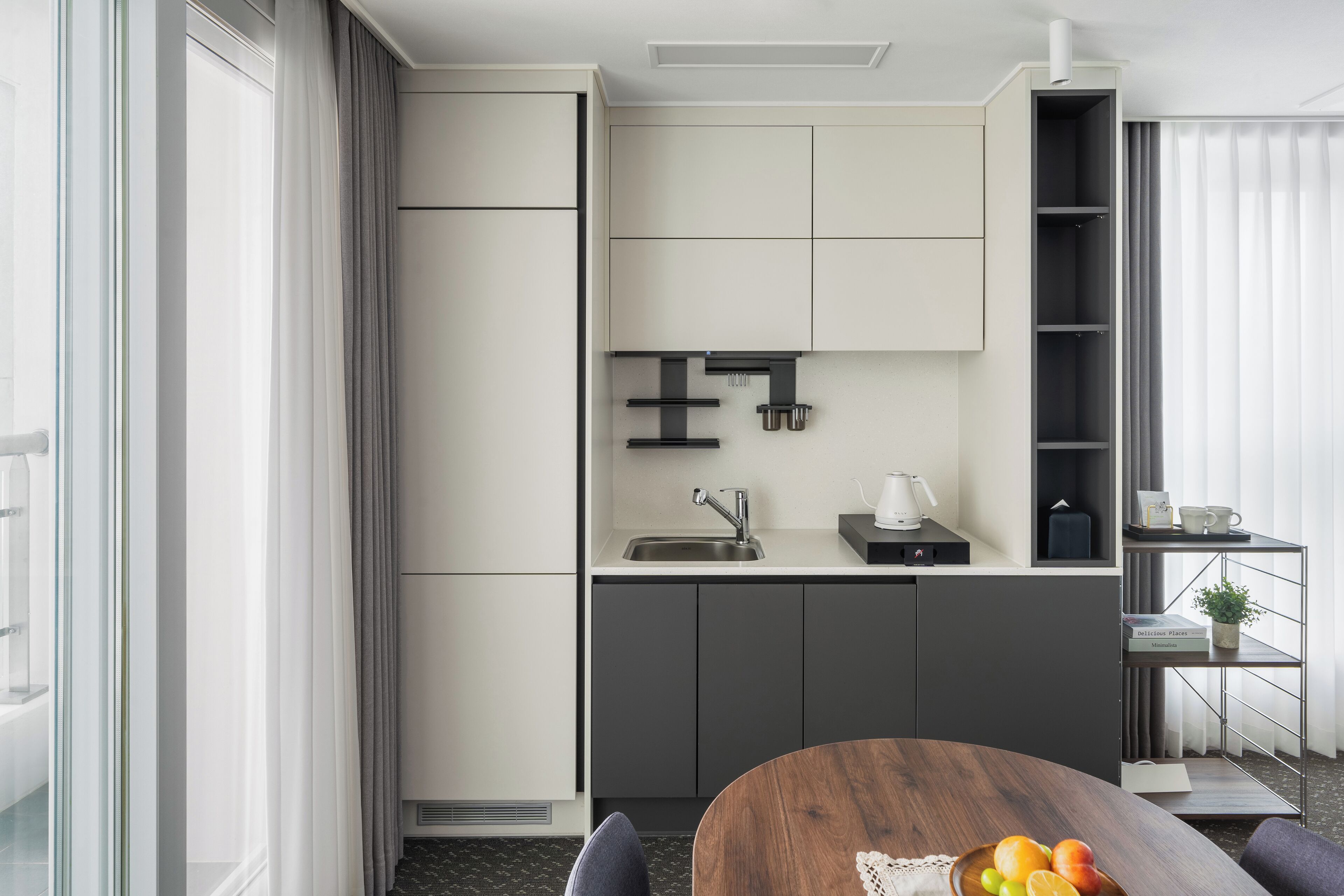 A modern kitchen with a built-in oven, a sink, and a coffee machine. There is a dining table with a fruit bowl.