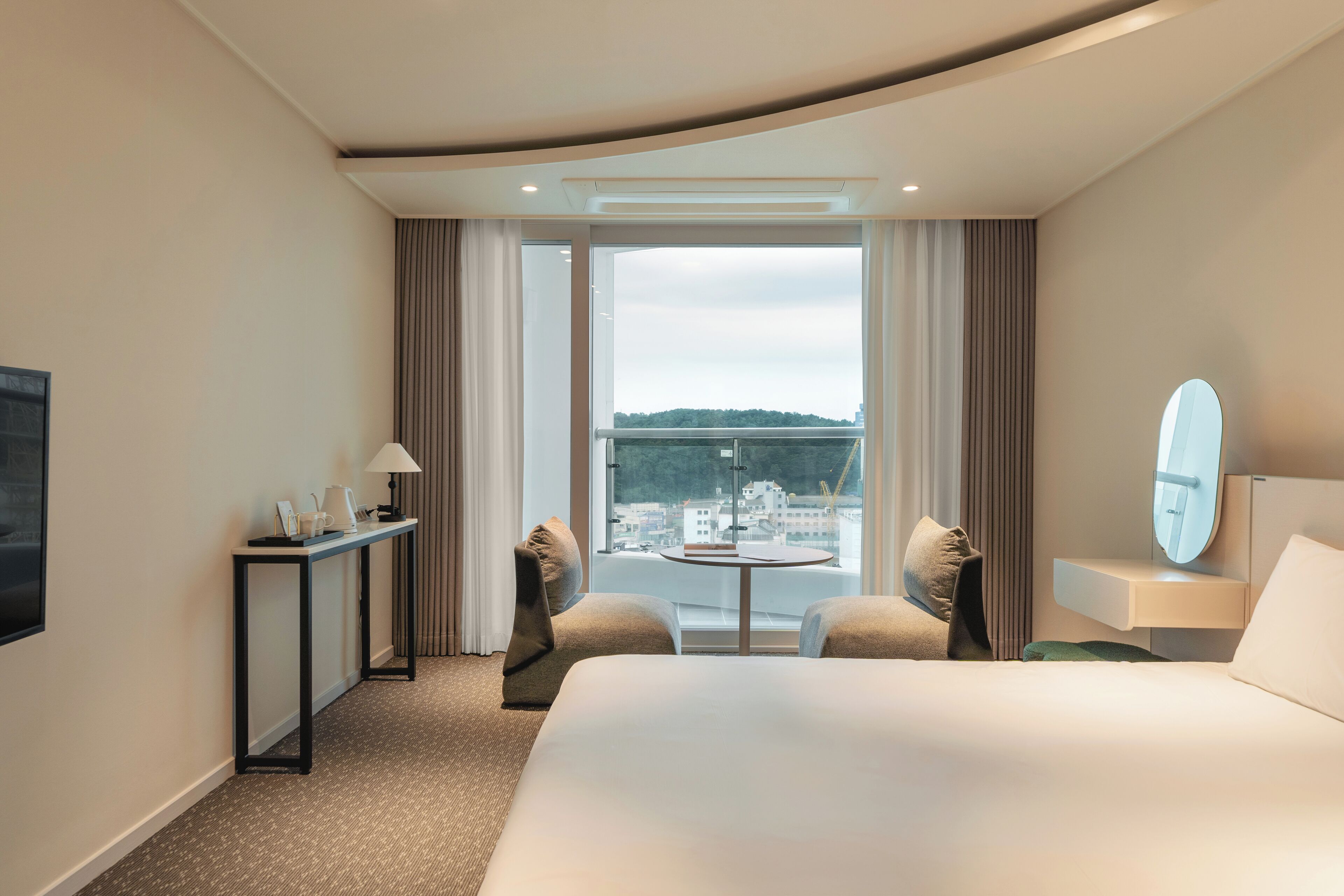 Quarto de hotel com uma cama grande, uma mesa pequena, duas poltronas, uma escrivaninha com luminária, um espelho e vista para a cidade através de amplas janelas.