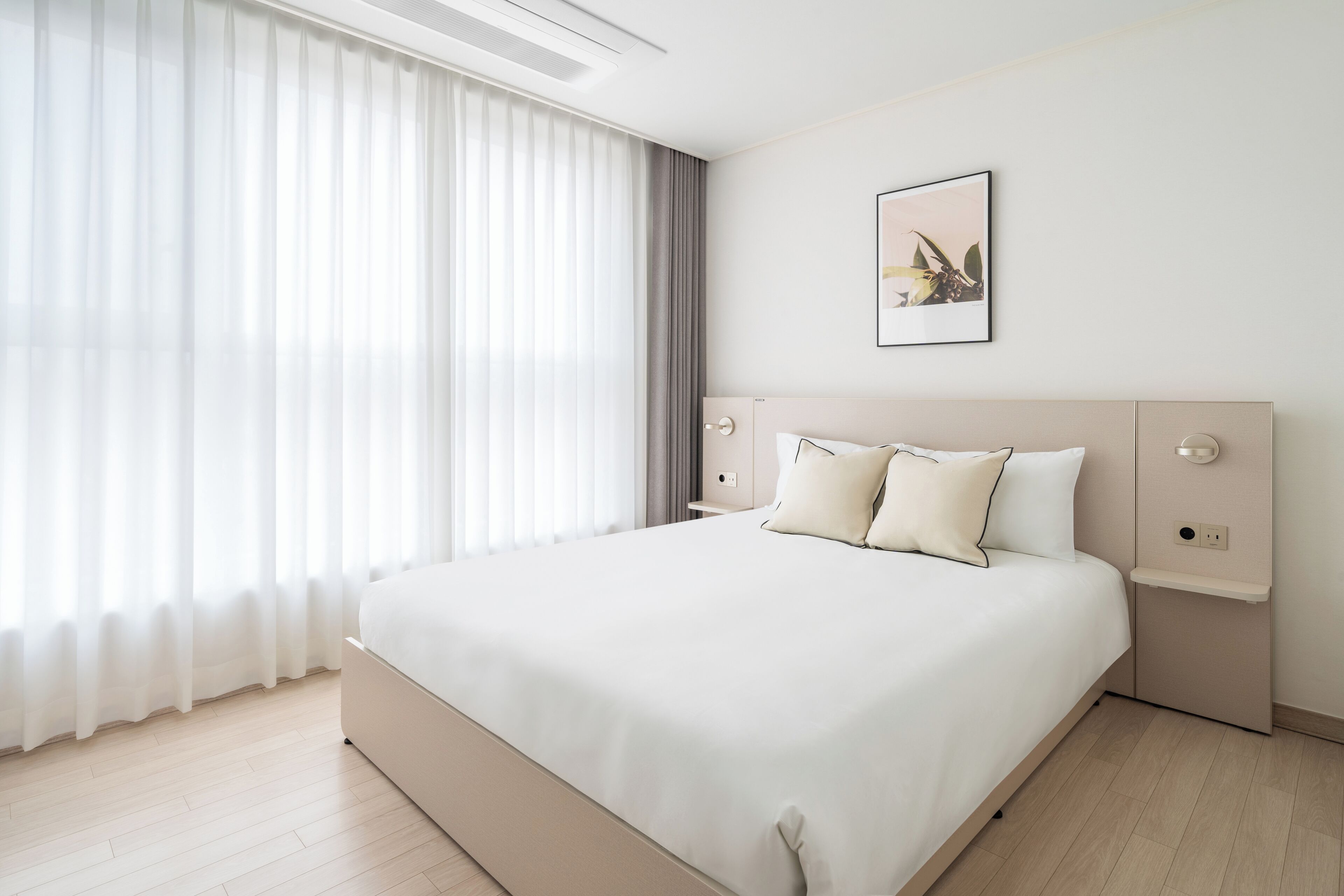 A modern hotel room with a large bed, a bedside table, a wall-mounted artwork, and sheer curtains.