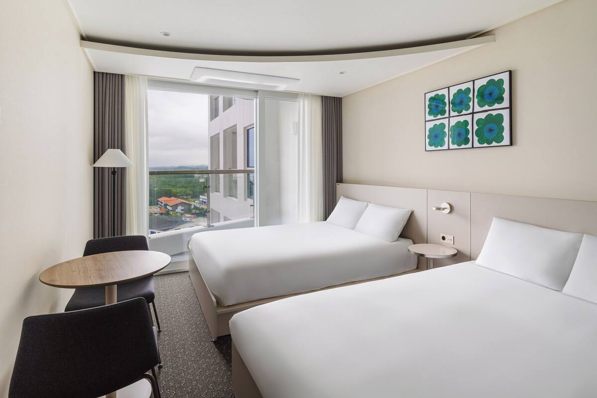 A hotel room with two beds, a small round table, a chair, a lamp, and a window with a view of buildings.