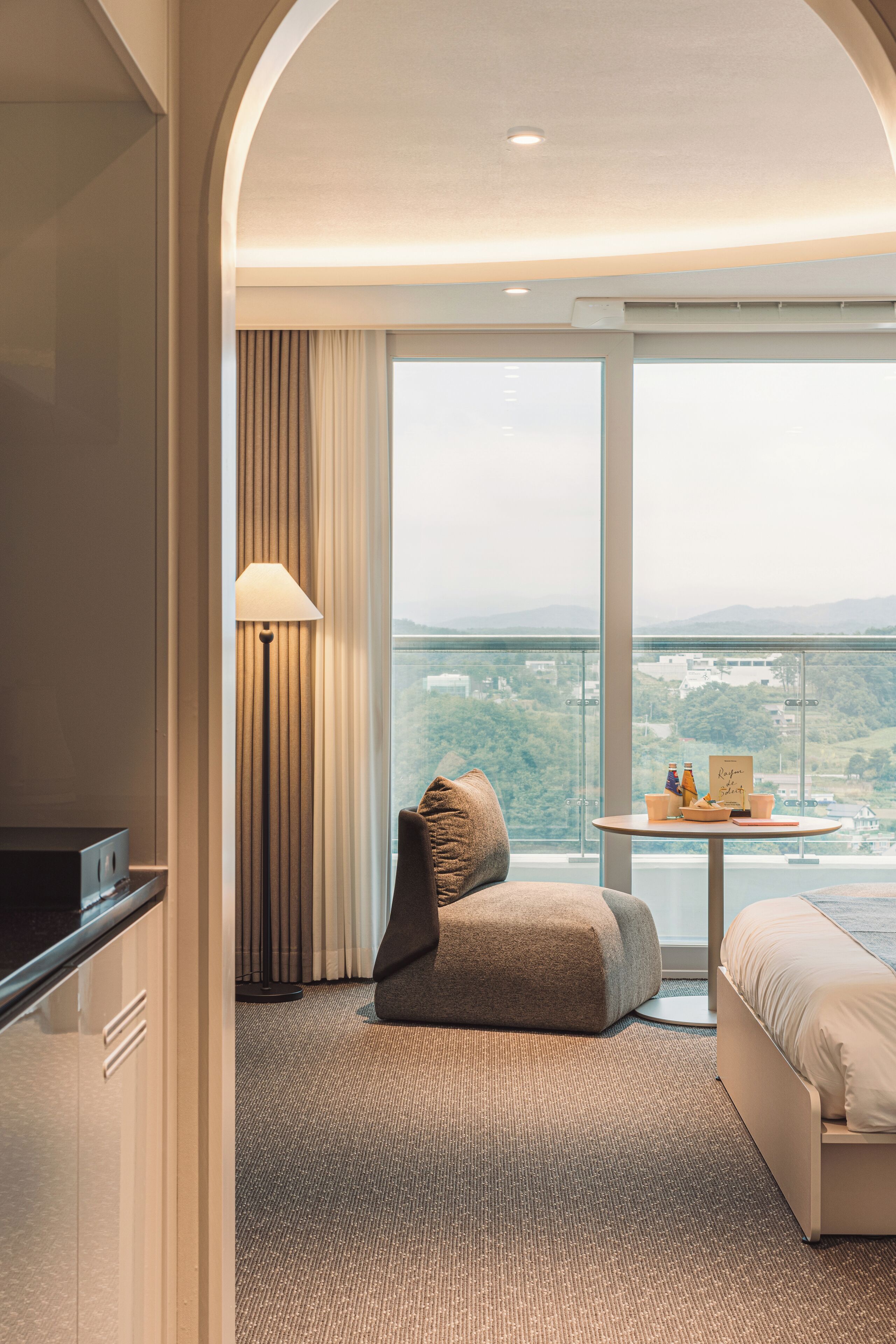 A modern hotel room with a large window, a sofa, a small table with a lamp, and a view of the city.