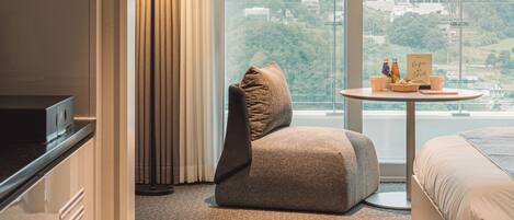 A modern hotel room with a large window, a sofa, a small table with a lamp, and a view of the city.