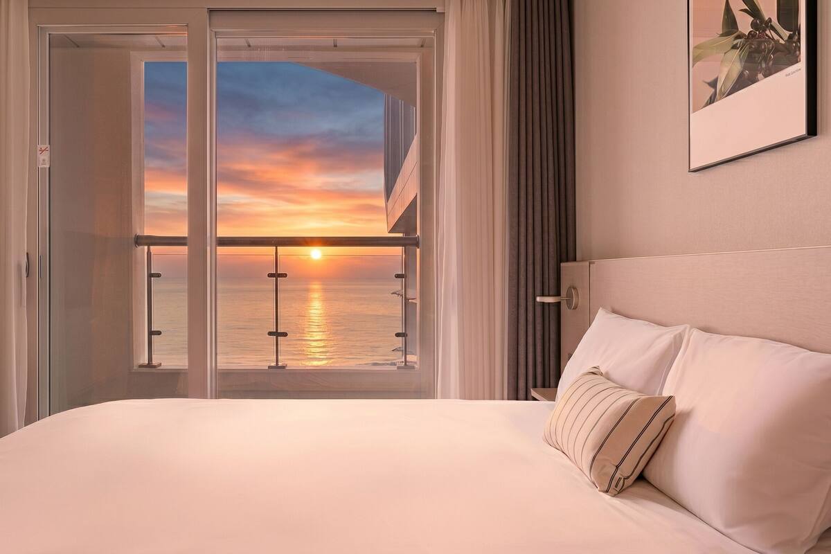 A hotel room with a bed, a view of the sunset over the water, and a balcony with a railing.