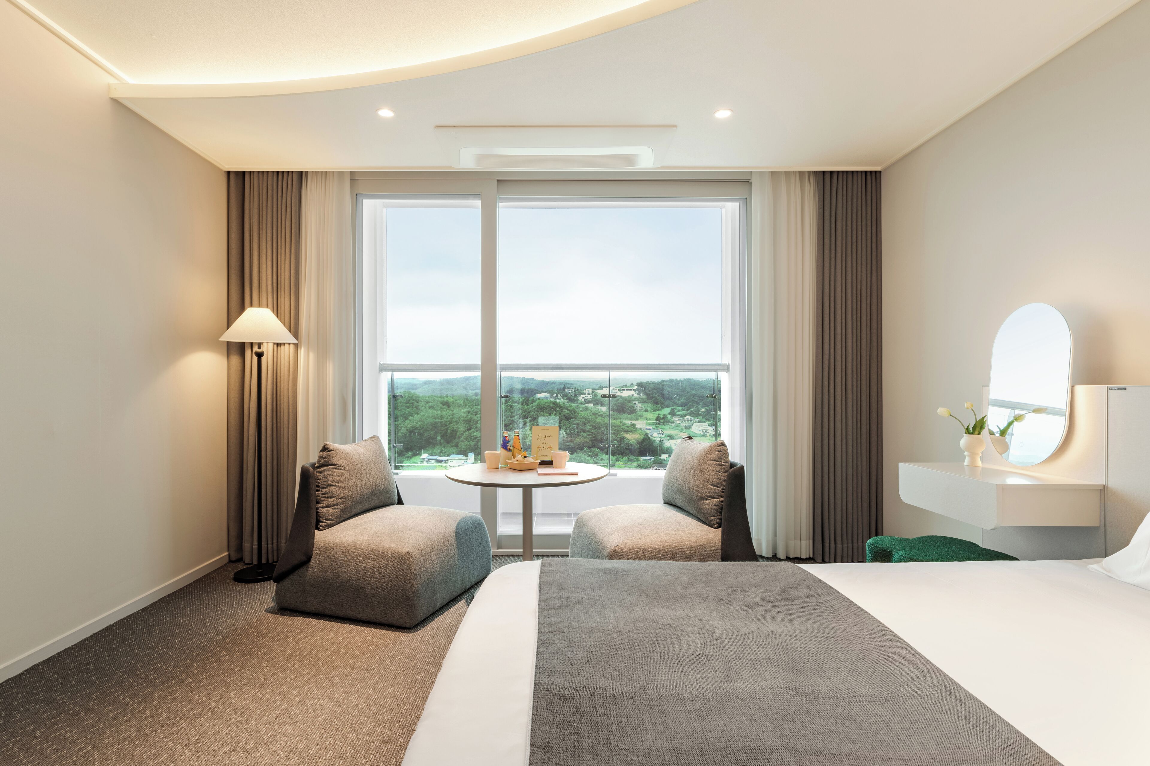 A modern hotel room with a large bed, two armchairs, a small round table with a view, and a large window.