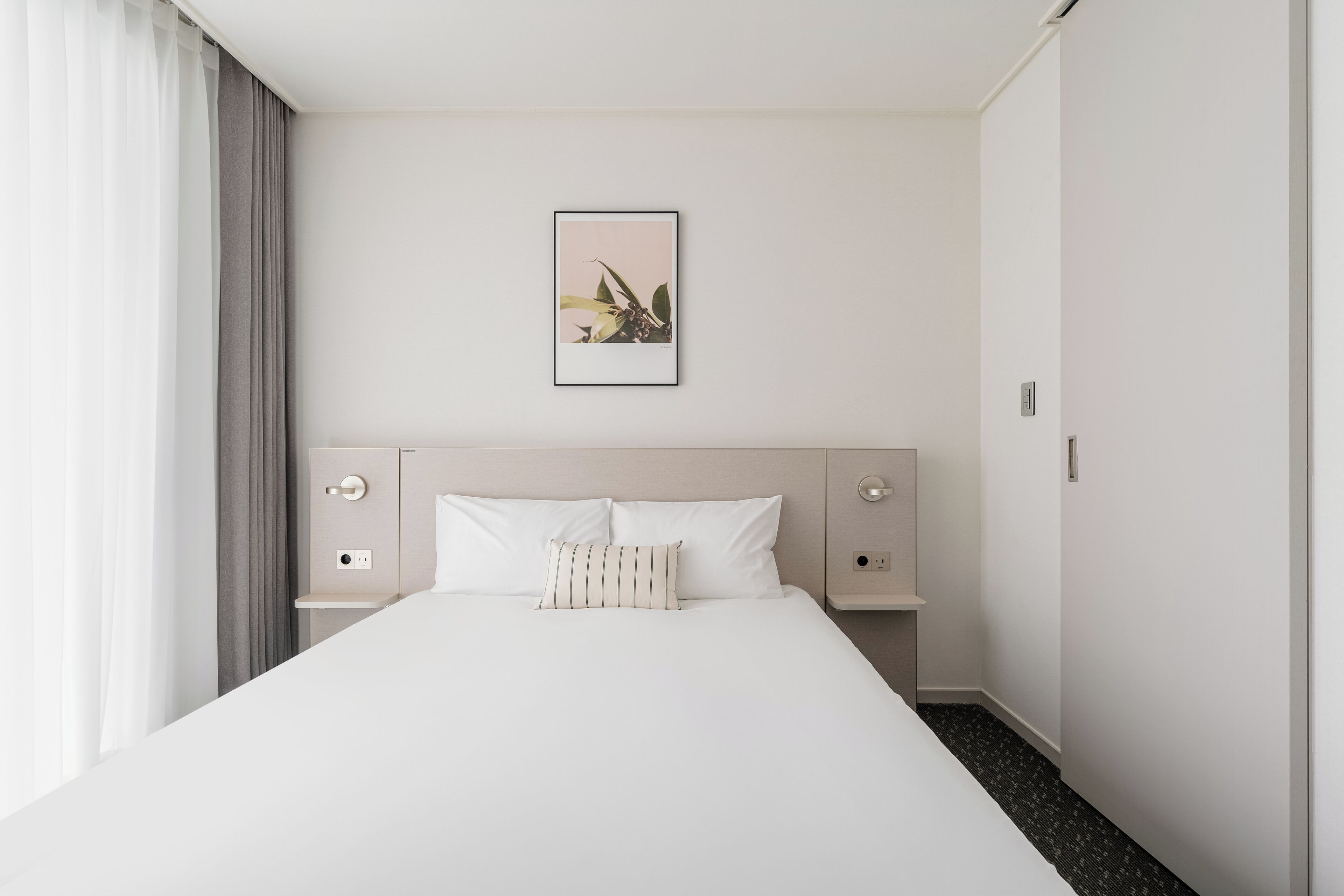A hotel room with a large bed, a bedside table, a nightstand, a framed picture on the wall, and a window with curtains.