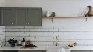 Una cucina con piastrelle bianche stile metro, mobili verdi, uno scaffale in legno con oggetti decorativi e un lavello con rubinetto dorato.