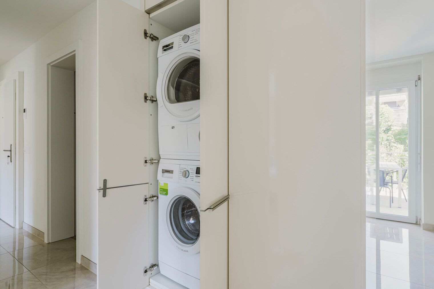 A laundry room with two stacked front-loading washing machines, a door leading to another room, and a view of the outside through a glass door.