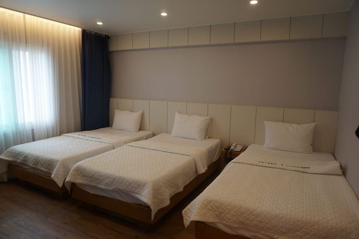 Ein Hotelzimmer mit drei Einzelbetten, einem Nachttisch mit Telefon und einem Fenster mit Vorhängen.