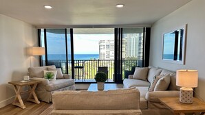 Ein Wohnzimmer mit zwei Sofas, einem Couchtisch und einem kleinen Beistelltisch mit Lampe. Große Fenster bieten Blick auf den Ozean und Gebäude.