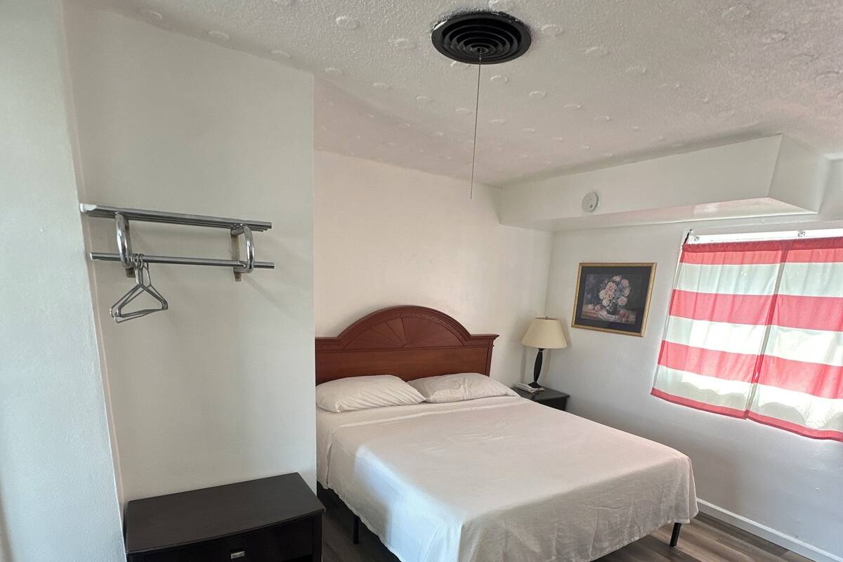 A bedroom with a bed, a bedside table, a lamp, a picture on the wall, and a window with curtains.