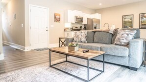 A modern living room with a grey sofa, a wooden coffee table, and a kitchen area with white cabinets and appliances. - Ducks Landing Accommodations at Southern Pines Golf Course (Aberdeen)