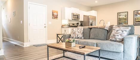 A modern living room with a grey sofa, a wooden coffee table, and a kitchen area with white cabinets and appliances.