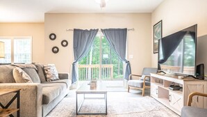 A living room with a sofa, coffee table, TV, and a balcony with curtains. - Ducks Landing Accommodations at Southern Pines Golf Course (Aberdeen)