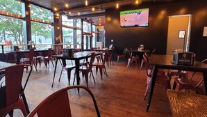 Un restaurante con mesas y sillas de madera, una pantalla grande mostrando un evento deportivo y amplias ventanas que dejan entrar luz natural.