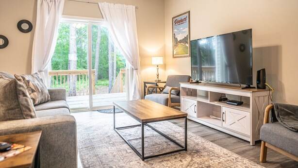 Una sala de estar con sofá, una mesa de centro, un televisor sobre un soporte de madera y un balcón con cortinas.