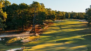 Un campo de golf con un camino despejado, búnkeres de arena y  árboles al fondo.