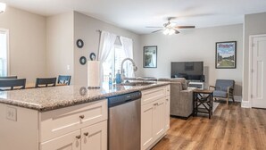 A modern kitchen with white cabinets, a granite countertop, and a dishwasher. There is a dining area with a table and chairs, and a living room with a sofa and a television. - Ducks Landing Accommodations at Southern Pines Golf Course (Aberdeen)