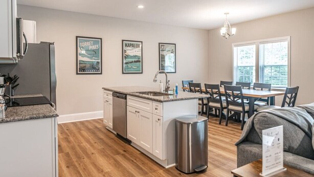 A modern kitchen with white cabinets, a granite countertop, and a stainless steel sink. There are framed posters on the wall and a dining area with a table and chairs. - Ducks Landing Accommodations at Southern Pines Golf Course (Aberdeen)