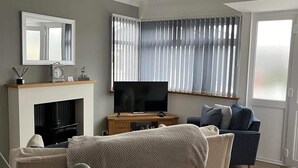 A living room with a sofa, TV, fireplace, and a large window with blinds. - SeasideEscape-2Br-Sleeps4-Parking-PetsOK-Fireplace (Bridlington)