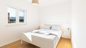 Een lichte slaapkamer met een groot raam, een bed met wit beddengoed, een nachtkastje en een hanglamp.
