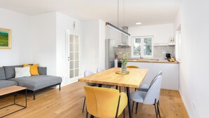 Un espacio habitable moderno con comedor, un sofá gris con cojines amarillos, una mesa de comedor de madera y una cocina al fondo.