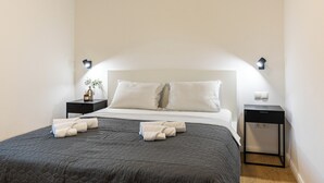 Una cama bien tendida con un edredón oscuro, dos almohadas y toallas dobladas sobre un cabecero blanco. Hay dos mesitas de noche, una con una lámpara y la otra con un enchufe.
