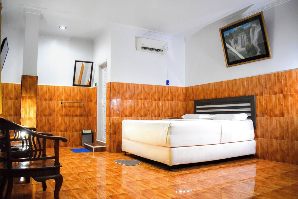 A hotel room with a wooden floor, a bed with white bedding, a wooden headboard, a wooden bench, a wall-mounted air conditioner, and a framed picture of a waterfall.