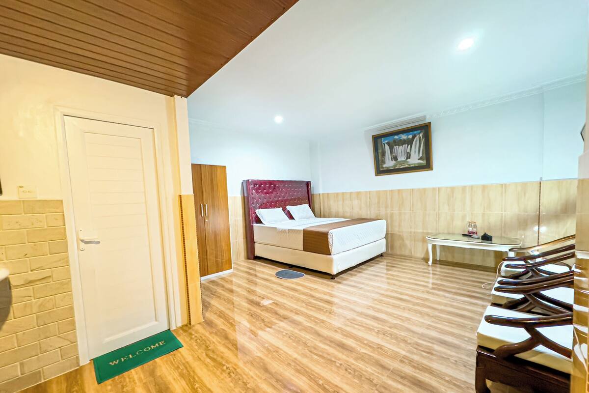 A hotel room with a wooden floor, a bed with a red headboard, a small table, and a chair.
