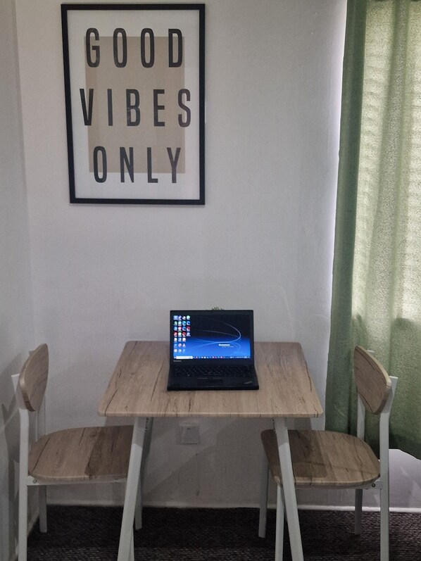 A laptop on a wooden table with two chairs, a framed sign saying "GOOD VIBES ONLY" on the wall, and a green curtain partially visible.