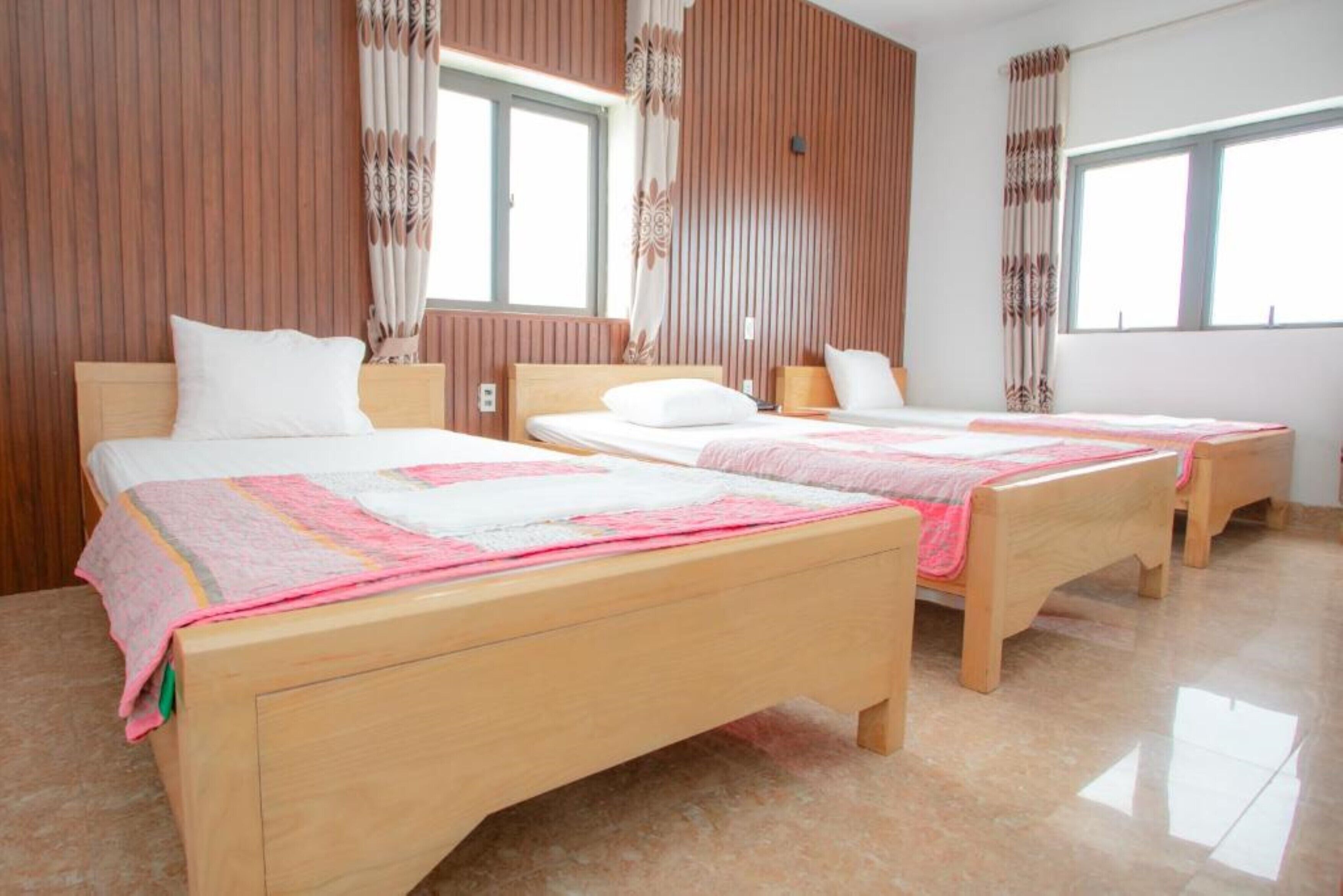 Een hotelkamer met twee houten bedden, elk voorzien van een wit kussen en een roze deken. De kamer heeft houten lambrisering en grote ramen met gordijnen.