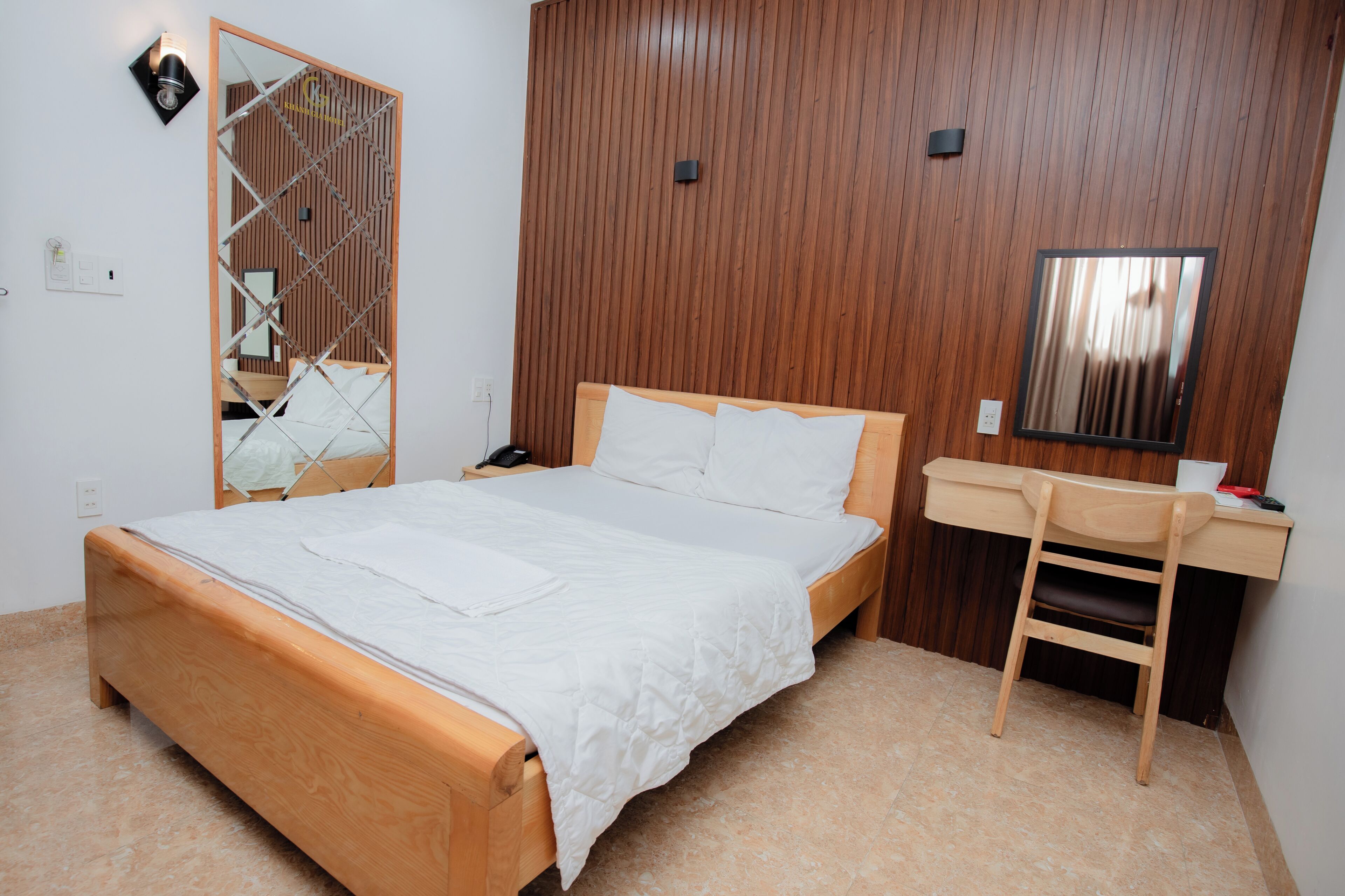A hotel room with a wooden bed, a desk with a chair, a mirror, and a wall-mounted light fixture.