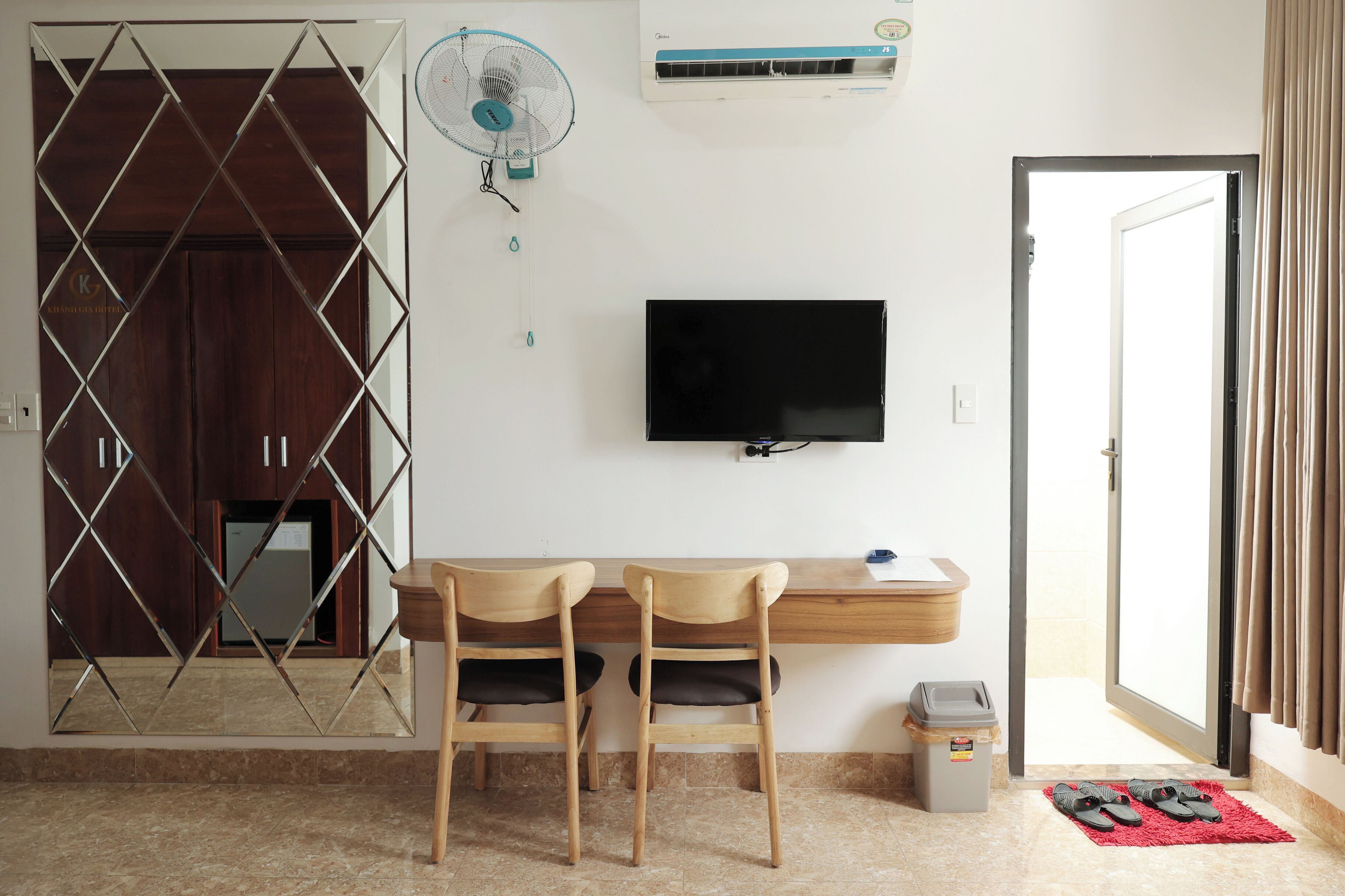 A hotel room with a desk, two chairs, a TV, a fan, and a door.