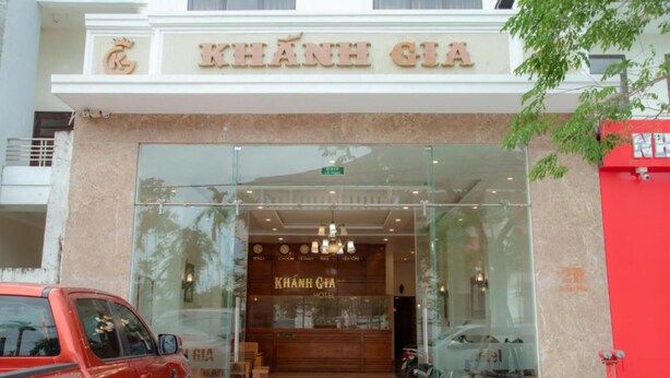 L’entrée de l’hôtel Khánh Gia, avec sa façade en verre, une enseigne au-dessus de l’entrée et un véhicule rouge garé à l’extérieur.