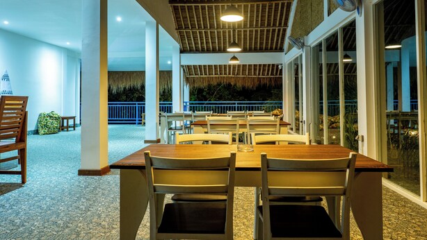 A spacious indoor dining area with a long wooden table and chairs, large windows, and a ceiling with a woven pattern. - Turtle House Lombok (Kuta)