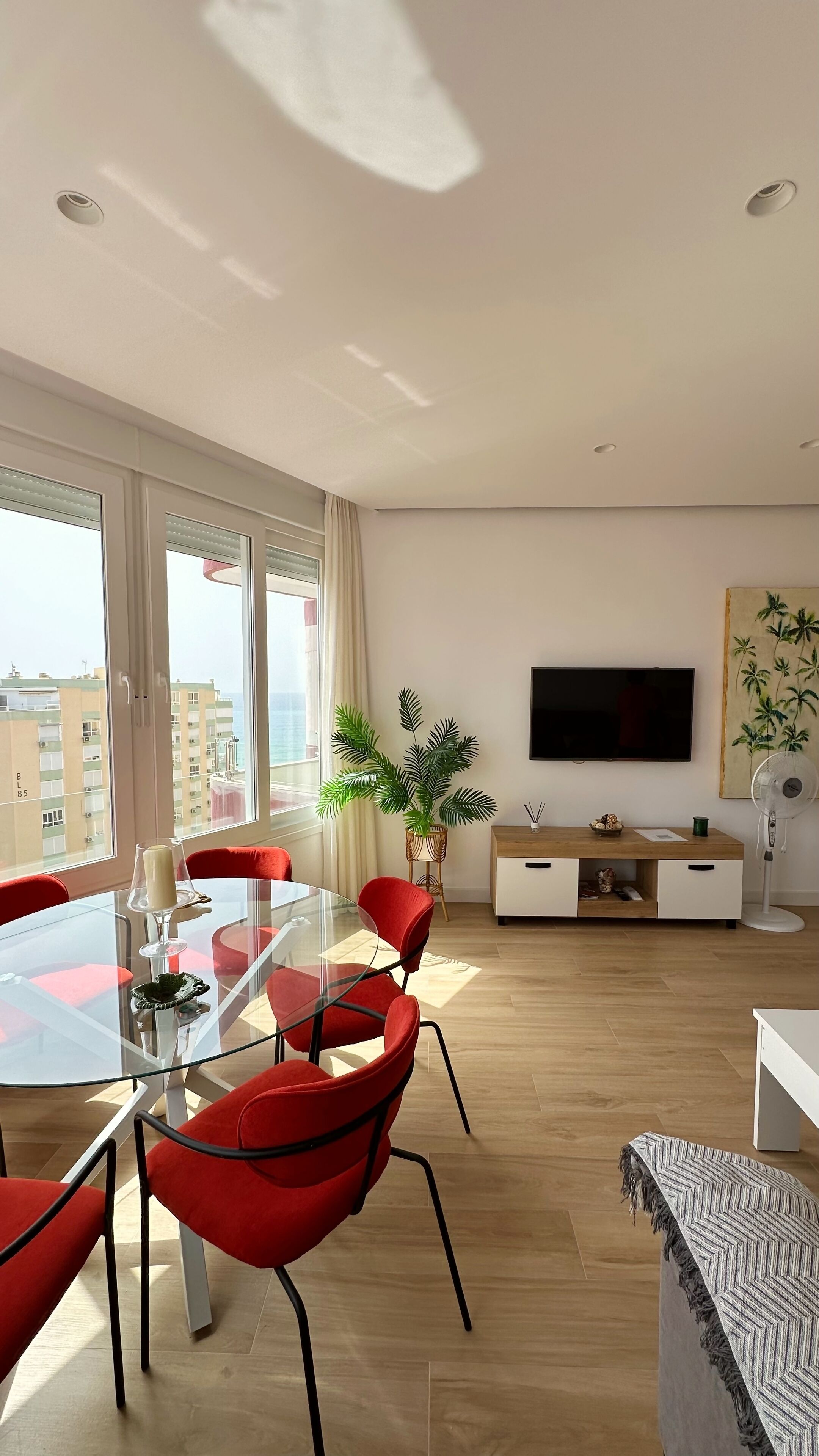Un salon moderne avec une table à manger en verre, des chaises rouges, une grande fenêtre donnant sur des immeubles, un téléviseur à écran plat et une plante en pot.