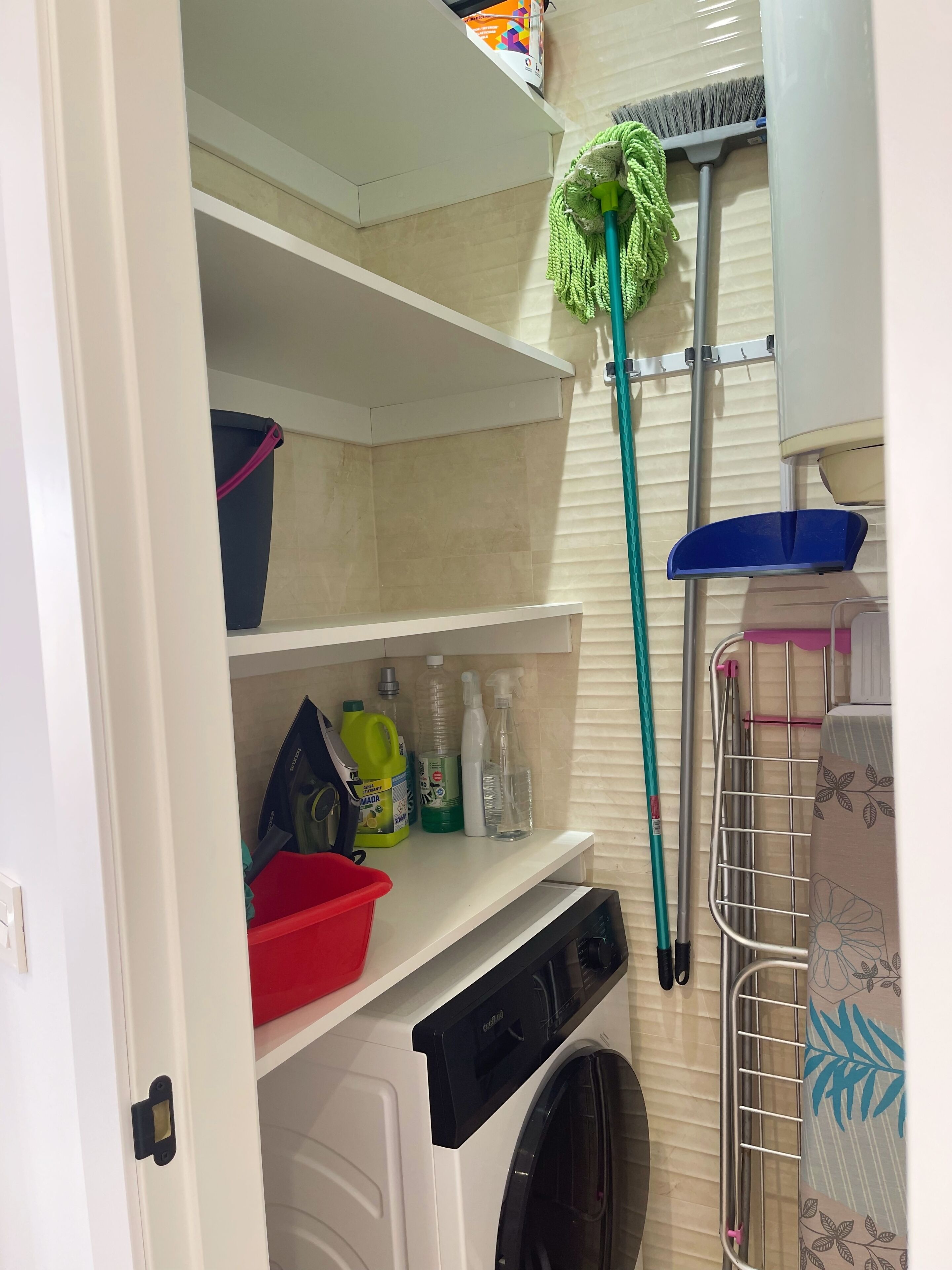 A laundry area with a washing machine, shelves with cleaning supplies, a mop, and a drying rack.