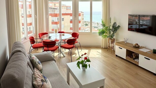 A modern living room with a sofa, a coffee table, and a TV. There are large windows with a view of buildings and a balcony. - DELUX Penthouse International Center (Llanos, Los (Torrox-Costa))