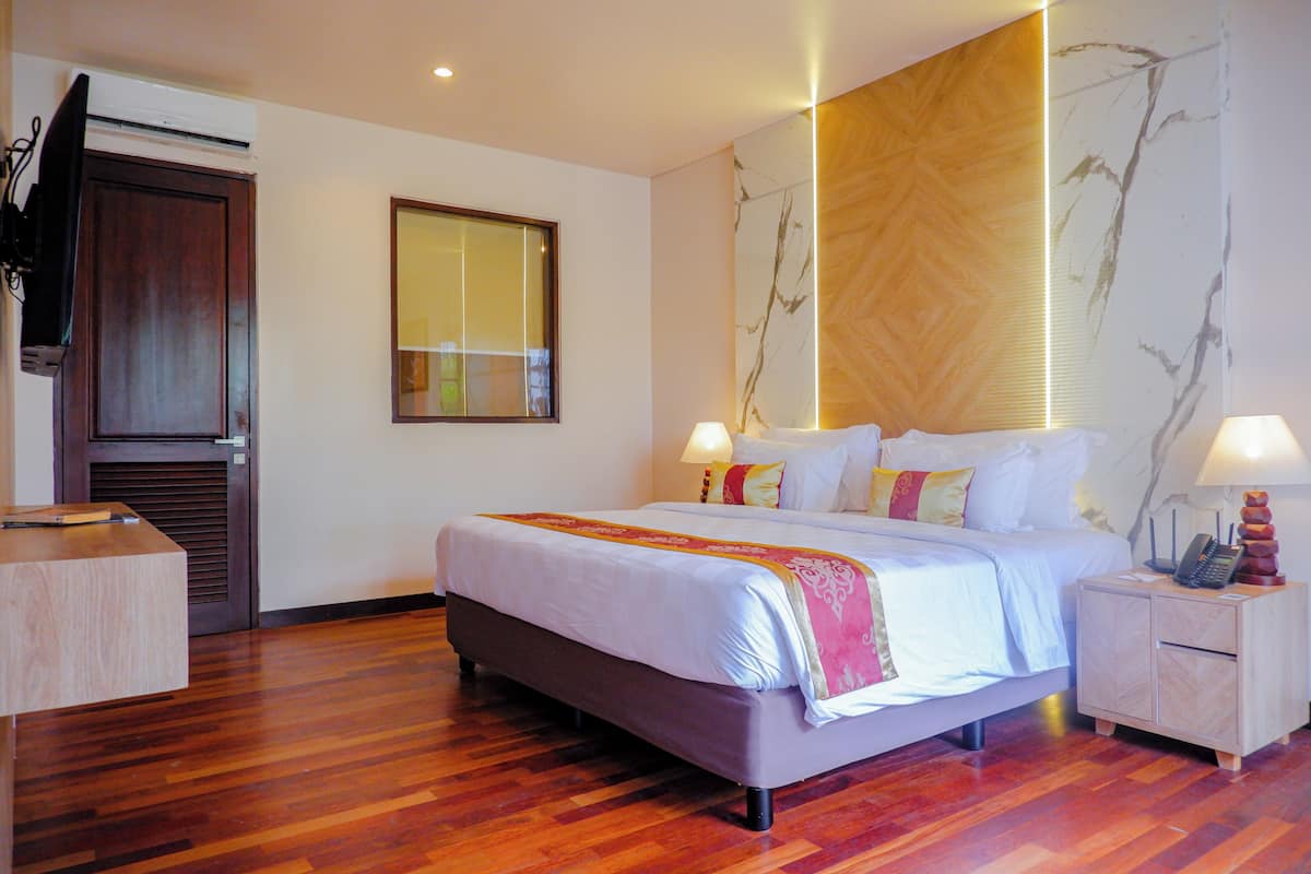Een hotelkamer met een houten vloer, een bed met witte lakens en een roze plaid, een nachtkastje met een lamp, en een aan de muur bevestigde televisie.