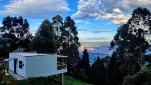 A modern white building with a balcony, surrounded by lush greenery and tall trees, overlooking a scenic landscape. - Cabaña en la Montaña con Vista a la Ciudad Desde el Jacuzzi (Medellín)