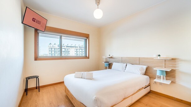 A modern bedroom with a large bed, wooden headboard, a small stool, and a window with a city view. - GuestReady - Hideaway in Porto (Porto)