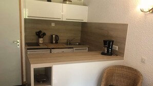 Kompakti keittiö, jossa on valkoiset kaapit, puinen työtaso, kahvinkeitin ja rottinkituoli, jossa on pehmuste.