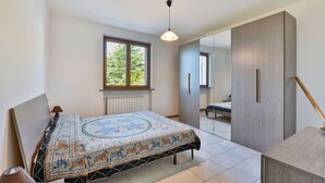 Un dormitorio con una cama grande, un radiador, una ventana que ofrece vistas a árboles, un armario y una puerta que da a otra habitación.
