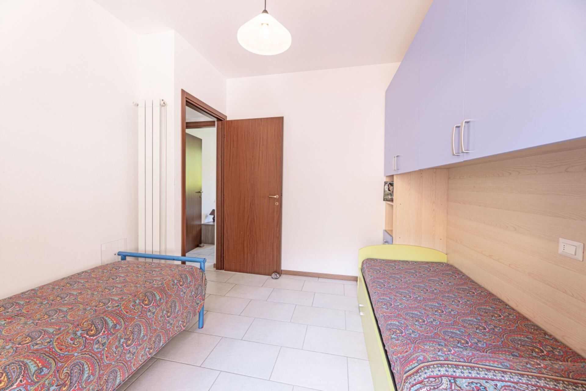 A bedroom with two single beds, a wooden wardrobe, a door leading to another room, and a light fixture on the ceiling.