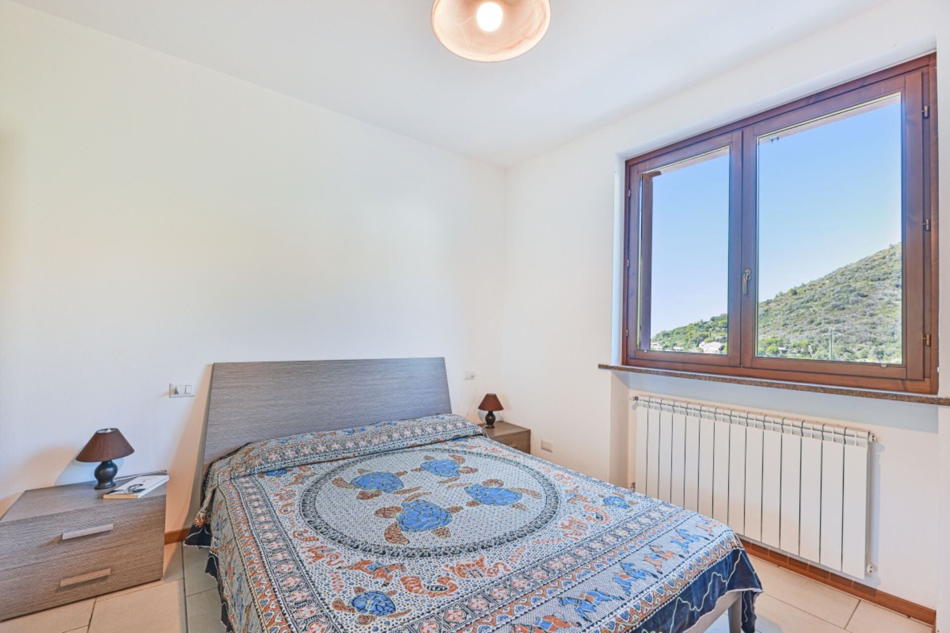 A bedroom with a bed, a nightstand, a lamp, a window with a view of a hill, and a radiator.