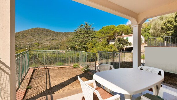 Un patio cubierto con una mesa y sillas blancas, una valla metálica y vistas a una colina con árboles y edificios.